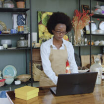 A small-business owner works on a laptop in a shop filled with handmade goods.