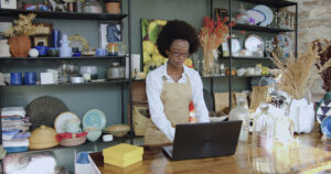 A small-business owner works on a laptop in a shop filled with handmade goods.