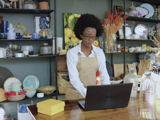 A small-business owner works on a laptop in a shop filled with handmade goods.