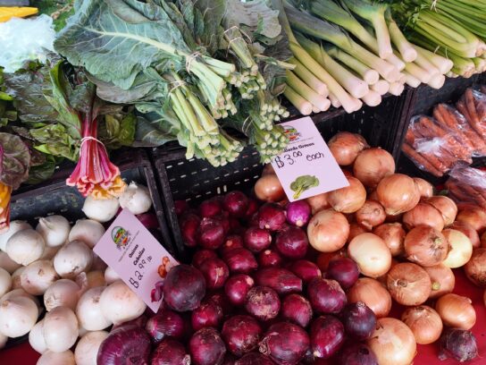 Bins of red, white and yellow onions, collard greens, chard, leeks and carrots at a Crossroads Farmers Market vendor stand, with a hand-written price sign reading "Onions Cebollas $2.99 lb."