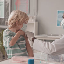 A doctor administers a vaccine to a young boy's arm while both wear face masks in a medical office.