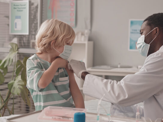 A doctor administers a vaccine to a young boy's arm while both wear face masks in a medical office.