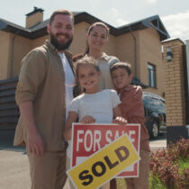 Family standing in front of a new home, posing with "Sign for Sale" and "Sold" sticker on it