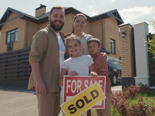 Family standing in front of a new home, posing with "Sign for Sale" and "Sold" sticker on it